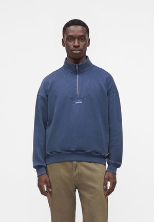 Mann trägt einen blauen Viertel-Reißverschluss-Sweatshirt mit Logo und khakifarbene Hose, steht vor einem schlichten weißen Hintergrund.
