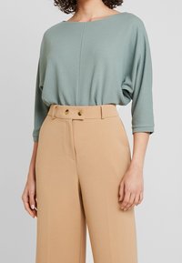 Kvinna i en strukturerad ljusgrön blus med trekvartsärmar, instoppad i högmidjade beige byxor med dubbelknappstängning.