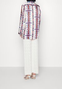 Chemise à manches longues en tissu à motifs avec des formes géométriques rouges et bleues, associée à un pantalon large blanc au motif chevron.
