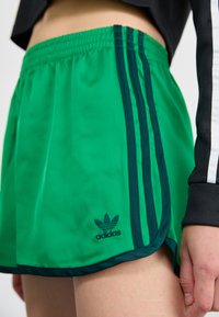 Green athletic shorts made of lightweight fabric, featuring three dark green stripes on the side and a small black Adidas logo.