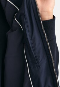 Navy jacket interior with a smooth, textured fabric, featuring a contrasting white piping and a zippered pocket along the seam.