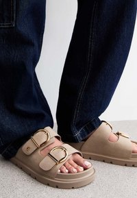 Beiga sandaler med två stora gyllene spännen, mjukt material och öppen tå-design, kombinerade med mörka denimjeans. Detaljerad sömnad synlig.