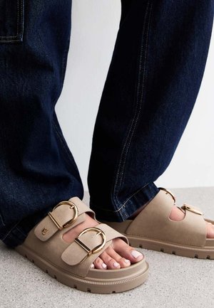Beiga sandaler med två stora gyllene spännen, mjukt material och öppen tå-design, kombinerade med mörka denimjeans. Detaljerad sömnad synlig.