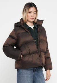 Hnědá péřová bunda s kapucí, s prošívaným designem, lesklou tkaninou a zipovým zapínáním. Nosič má na sobě zelenou kordovou košili.