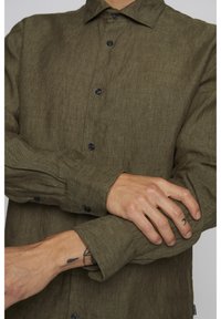 Olivno zelena lanena srajca z gumbom na sprednji strani, dolgimi rokavi in črnimi gumbi. Ima teksturirano tkanino in kontrastne manšete.