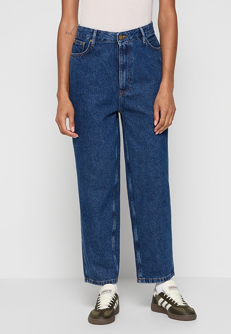 Person, der hochtaillierte, gerade geschnittene, blaue Jeans mit Knopf und Reißverschluss trägt, kombiniert mit weißen Socken und schwarz-weißen Adidas-Sneakern.