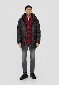 Zwarte pufferjas met capuchon, voorzien van een ritssluiting en knoopsluiting, met twee voorzakken, gedragen over een rood geruit overhemd en grijze jeans. Zwarte sneakers.