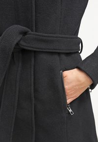 Black wool coat with a belted waist, featuring a textured fabric. Side pockets have zippers, and the design includes long sleeves and a tailored fit.