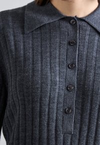 Dunkelgrauer ribbeliger Strickpullover mit weitem Kragen und fünf schwarzen Knöpfen auf der Vorderseite, getragen von einer Person.