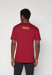 Burgundi värvi puuvillane T-särk, millel on ümar kaelus ja lühikesed varrukad. Seljaosas on sõna "BARÇA" julgete kuldsete tähtedega.