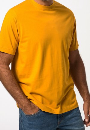 Homme portant un t-shirt jaune uni à manches courtes avec un jean bleu, une main dans la poche, sur un fond blanc.