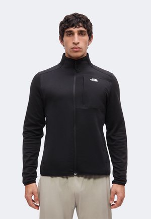 Muž nosiaci čiernu bundu na zips s vysokým golierom a logom The North Face na hrudi v kombinácii so svetlo béžovými nohavícami.