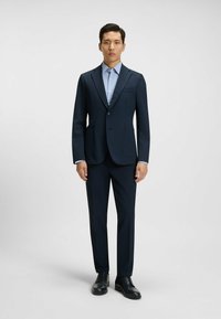 Completo blu scuro con una vestibilità slim, caratterizzato da una giacca a un bottone, due tasche frontali e una camicia a quadri blu sotto, abbinato a scarpe nere.