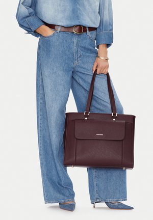 Person trägt lockere hellblaue Jeans und ein Jeanshemd, hält eine dunkelbraune Ledertasche mit doppeltem Griff.