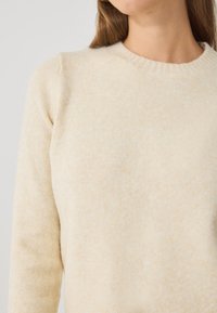 Suéter de un suave color beige claro, elaborado con una tela texturizada, que presenta un cuello redondo y mangas largas con un mínimo detalle en las costuras.