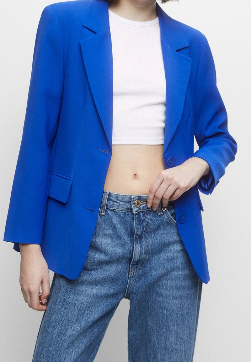 Personne portant un blazer bleu vif par-dessus un top court blanc et un jean bleu taille haute, une main touchant la ceinture du jean.