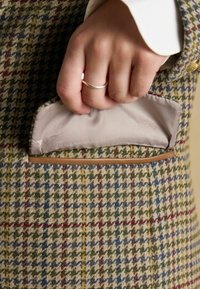 Vrecko na tweedovom blazeru v šedej, zelenej a červenej kocke, s hladkým, svetlo sfarbeným saténovým podšívkom a koženým lemovaním.