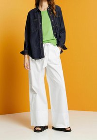 Femme portant un pull vert, une veste en denim foncé, un pantalon blanc à jambes larges avec ceinture à nouer, et des sandales noires sur un fond orange.