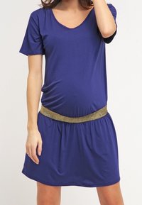 Violetti äitiysmekko, jossa on lyhyet hihat, pyöreä pääntie ja kerätty vyötärö. Kultaista kimaltelevat vyö korostaa suunnittelua.