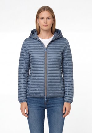 Vrouw die een lichtblauwe gesteepte jas met ritssluiting en capuchon draagt, samen met een blauwe spijkerbroek, staand tegen een witte achtergrond en vooruit kijkend.