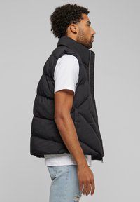 Schwarze Steppweste mit hohem Kragen, Reißverschluss und gestepptem Design. Kombiniert mit einem weißen T-Shirt und hellblauen Jeans.