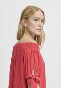 Top corallo off-the-shoulder con scollo elastico; maniche corte a sbuffo legate ai gomiti; tessuto con texture e delicate pieghe.