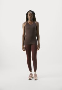 Hosszú copfokkal rendelkező nő áll szembe, barna trikót, bordó leggingset, fehér zoknit és rózsaszín sportcipőt viselve egy fehér háttér előtt.