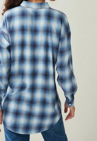 Camicia a quadri blu con maniche lunghe, caratterizzata da diverse tonalità di blu e bianco, orlo arrotondato e polsini abbottonati.