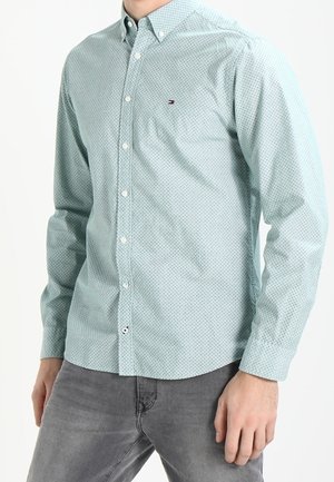 Hombre vistiendo una camisa de botones azul claro con estampado y jeans grises, de pie con una mano relajada a su lado.