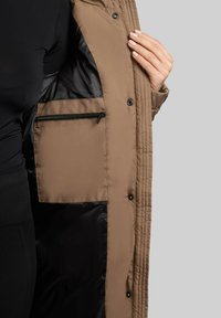 Abrigo acolchado marrón, exterior acolchado, cierres con botones y un bolsillo interior con cremallera negra. El forro es liso y oscuro.