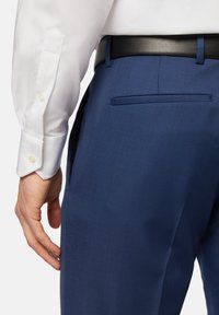 Marineblauwe broeken van een gestructureerde stof, met een achterzak, gecombineerd met een witte blouse met knoopmanchetten en een zwarte riem.