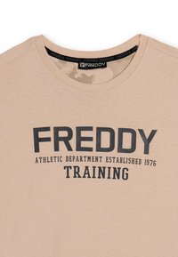 T-shirt en coton beige avec texte imprimé noir : "DÉPARTEMENT ATHLÉTIQUE FREDDY ÉTABLI EN 1976 ENTRAÎNEMENT." Design col rond. Texture lisse.