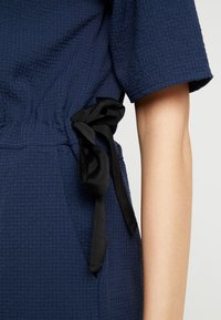 Marinblå texturerad klänning med korta ärmar, med en insvängd midja som är knuten med ett svart satinband. Synliga sidofickor.