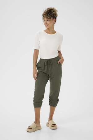 Weißes geripptes T-Shirt mit kurzen Ärmeln, olivgrüne Leinenhose mit elastischen Bündchen und Kordelzug, kombiniert mit beigen gewebten Slip-On-Sandalen.