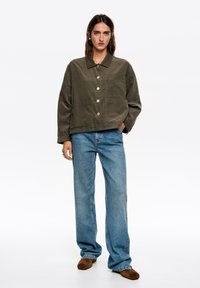 Khaki bawełniana kurtka z kołnierzykiem, z przednimi kieszeniami i dużymi guzikami, w zestawieniu z jasnoniebieskimi szerokimi dżinsami i brązowymi butami.