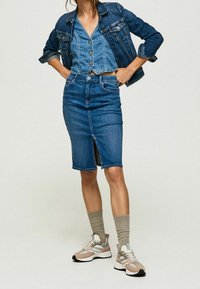 Kvinna som bär en blå denimjacka, knäppt blå denimväst, blå denimpennkjol med sprund fram, beige strumpor och sneakers, står med händerna på höfterna.