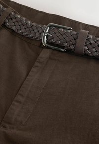 Ceinture en cuir tressé marron avec boucle argentée attachée à la taille d’un pantalon en coton marron foncé.