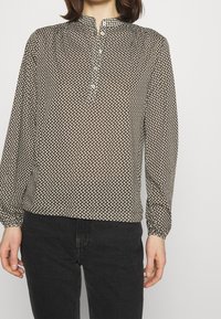 Braun gemusterte Bluse mit langen Ärmeln, Stehkragen und Knopfleiste. Besitzt ein kleines weißes Punktemuster auf einem transparenten Stoff.