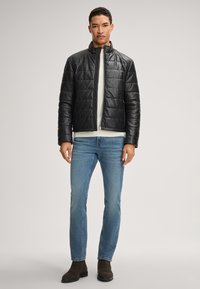 Schwarze gesteppte Lederjacke mit hohem Kragen, Frontreißverschluss, getragen über einem hellen Pullover und blauen Jeans, kombiniert mit braunen Stiefeletten.
