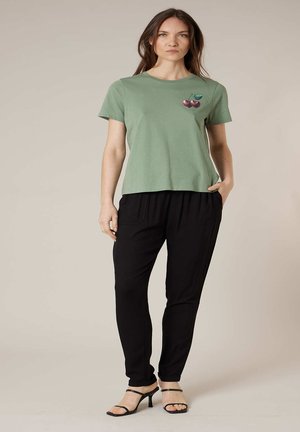 Femme portant un t-shirt vert clair avec un motif de cerise, un pantalon noir et des talons ouverts noirs, debout devant un fond uni.