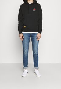 Svart huvtröja med känguruficka, prydd med röda, blå och orange Nike-logotyper. Bärs med ljusblå jeans och vita sneakers.