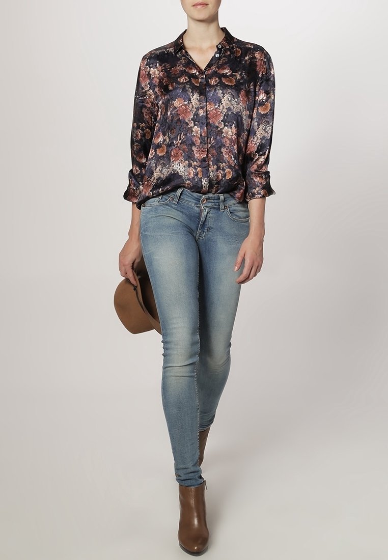 Personne portant un chemisier à fleurs foncé, un jean skinny bleu et des bottines marron, tenant un chapeau marron, debout devant un fond uni.