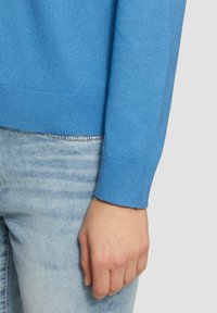 Blauer Strickpullover mit Rippenbund und -abschlüssen, der eine glatte Textur aufweist. Trägt dazu helle Denim-Jeans, und eine Hand ruht auf den Jeans.