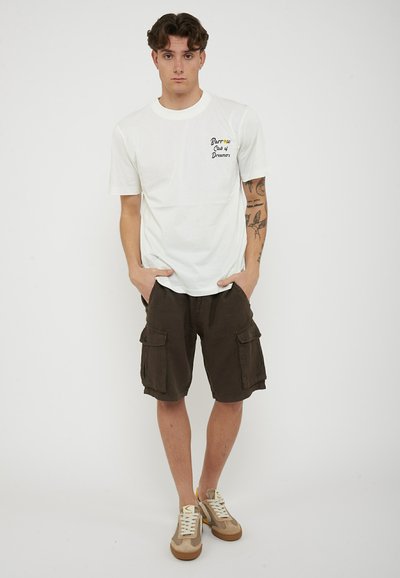 Giovane uomo in piedi con le mani in tasca, indossa una maglietta bianca "Barrow Club of Dreamers", pantaloncini cargo scuri, sneakers e tatuaggi visibili sulle braccia.