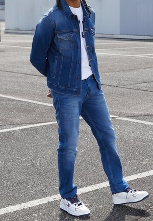 Person trägt blaue Jeansjacke und Jeans, weißes Hemd und weiße Sneaker, steht auf Asphalt mit Parklinien.