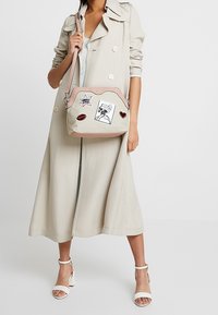 Femme portant un trench-coat beige et des talons blancs, tenant un sac bandoulière beige avec des bordures roses et des patchs graphiques sur le devant.