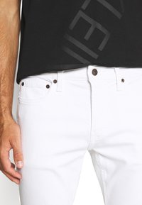 Valkoiset farkut slim fit -mallilla, joissa on ruskeat metalliset niitit, viisi taskua ja hillitty tekstuuri. Musta T-paita osittain näkyvissä yläosassa.