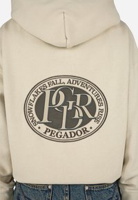 Sweatshirt bege com um grande gráfico circular nas costas, apresentando texto e logotipo em cinza escuro. Tecido de mistura de algodão com textura suave.