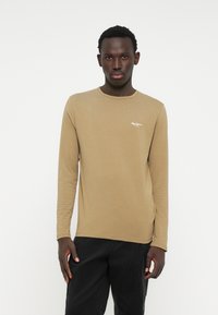 Maglietta a maniche lunghe color marrone chiaro in cotone, con scollatura a girocollo e un piccolo logo bianco stampato sul lato sinistro del petto.