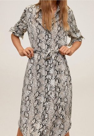 Femme portant une robe imprimée serpent, longueur genou, manches longues retroussées, attachant une ceinture à la taille sur un fond uni.
